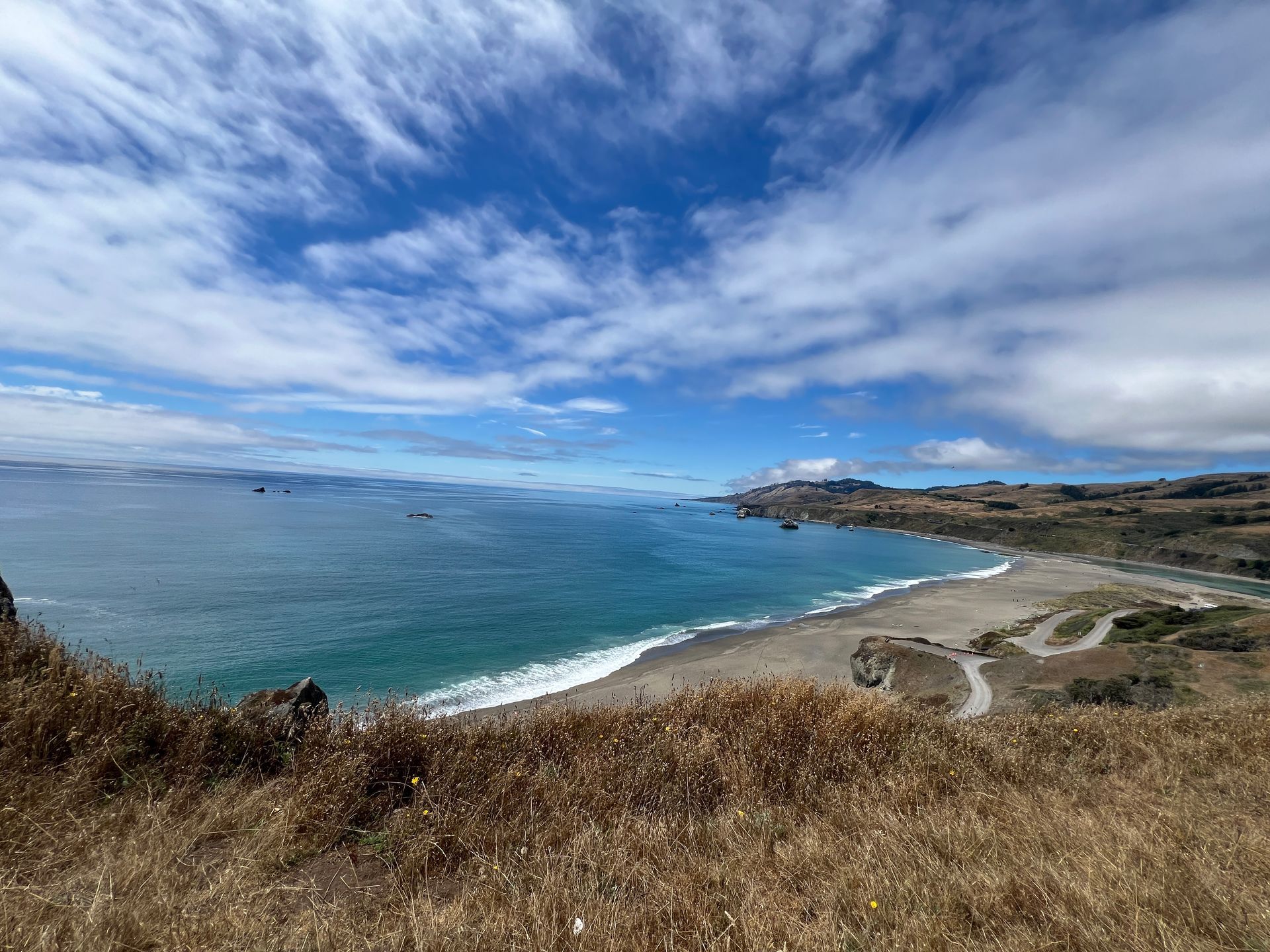 Coastal bay with blue ocean, sandy beach, and grassy foreground under a cloudy sky