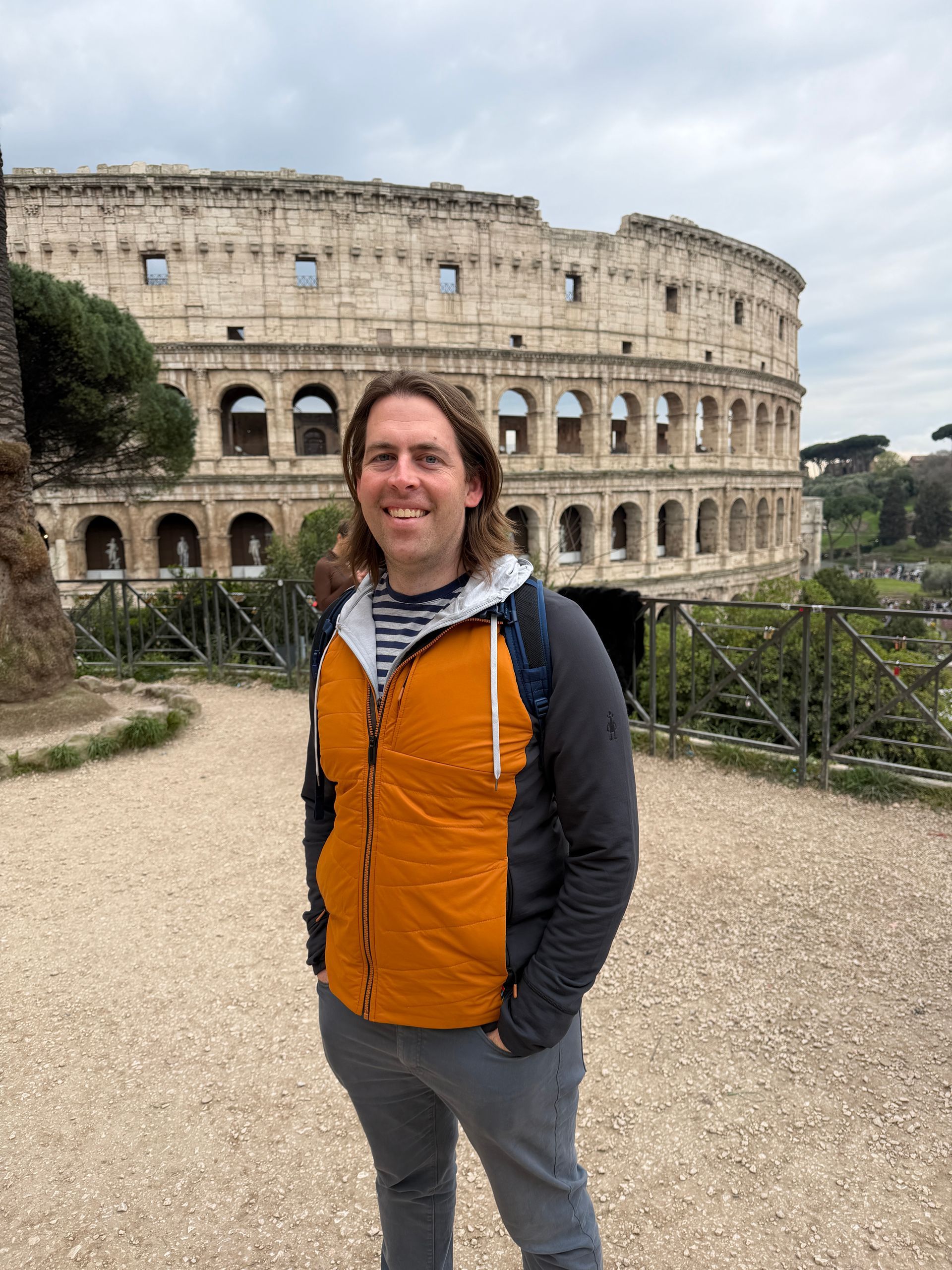 Brian Voss  standing in front of the Colosseum, wearing an orange jacket and backpack.