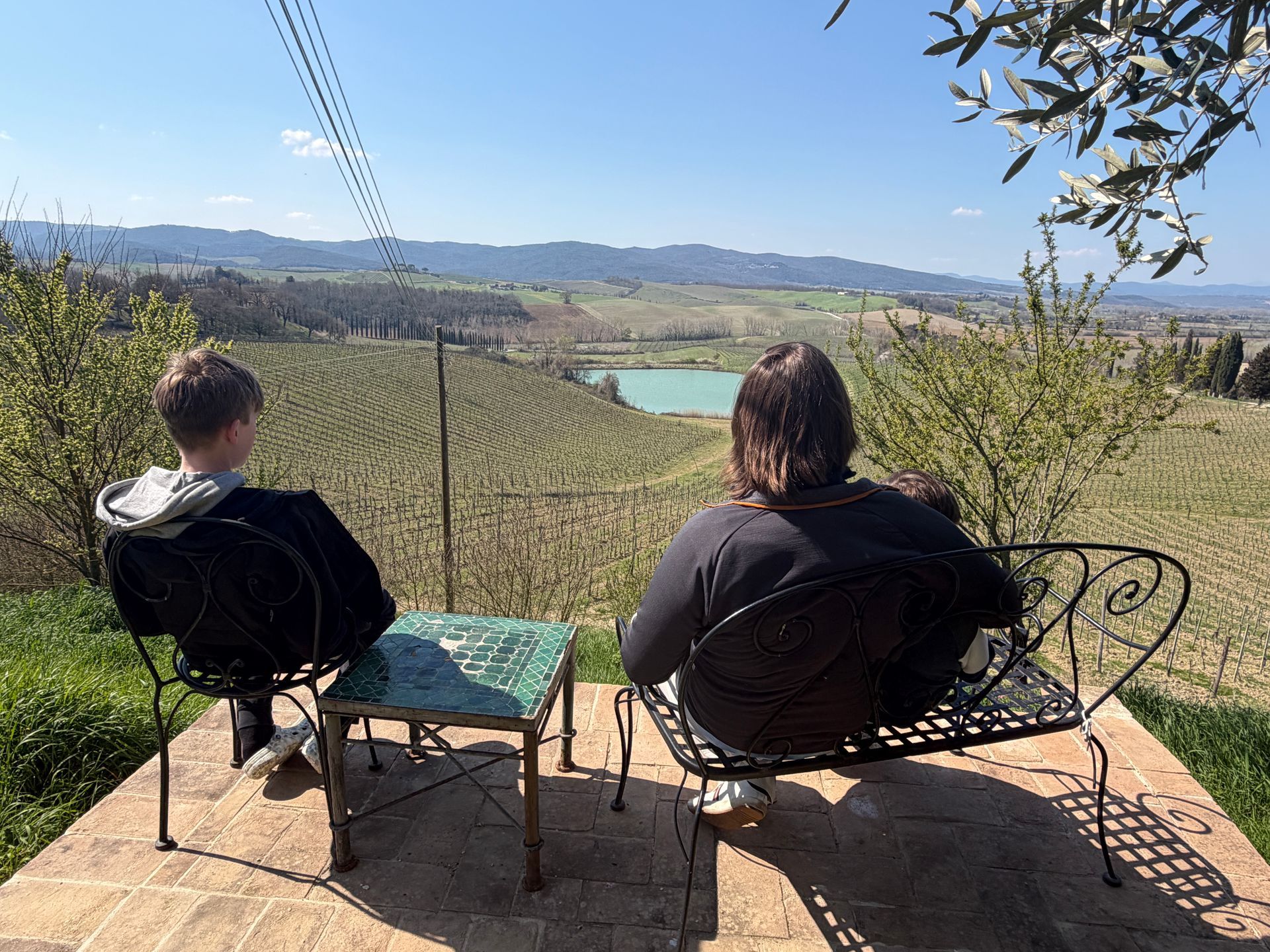 Two people sit on chairs overlooking a vineyard, lake, and mountains on a sunny terrace.