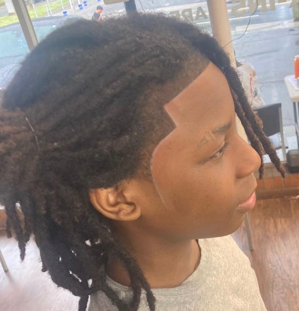 A young boy with dreadlocks and a shaved head is sitting in front of a window.