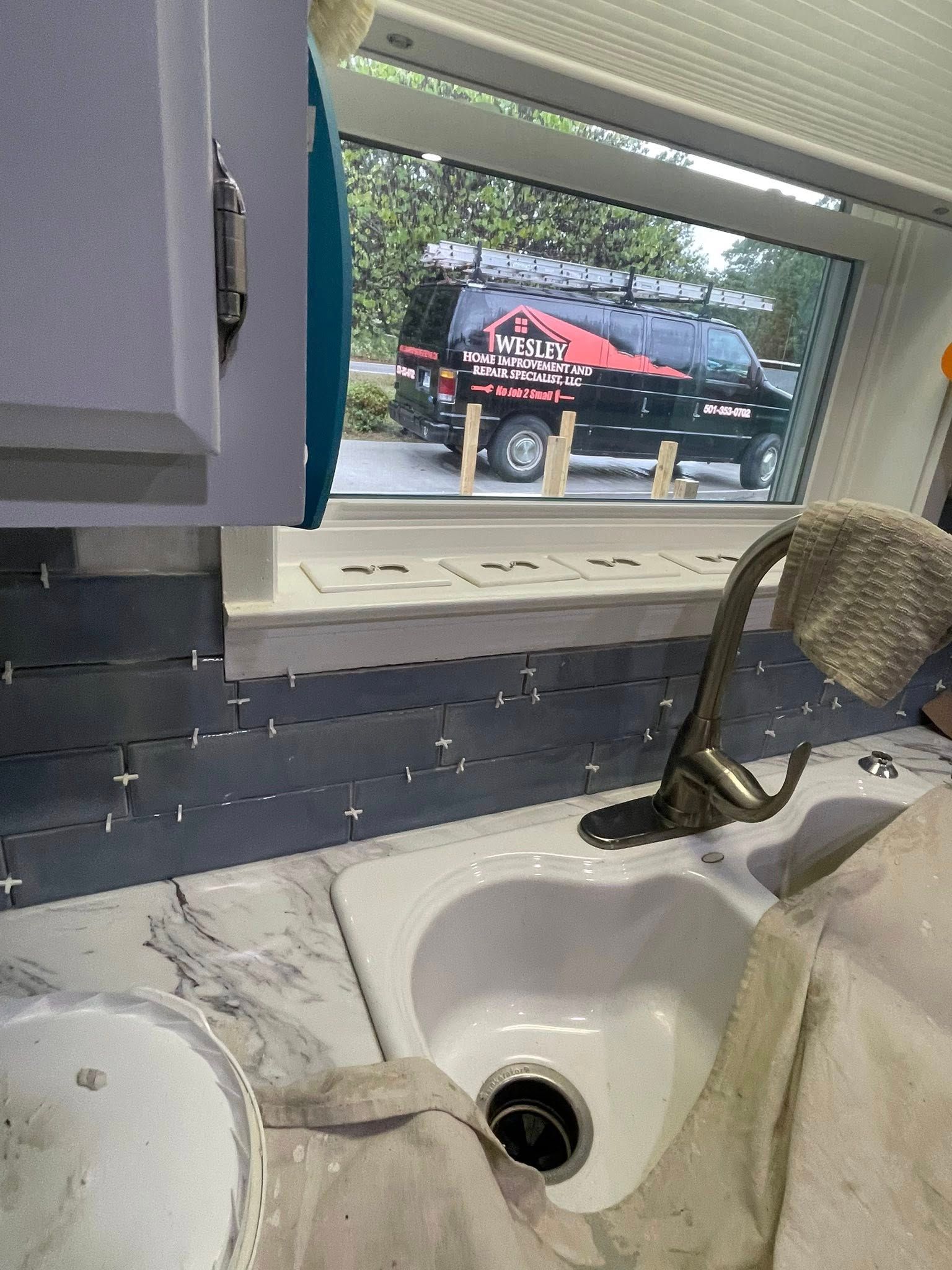 Kitchen interior with sink, tiling in progress, and work truck visible through a window.
