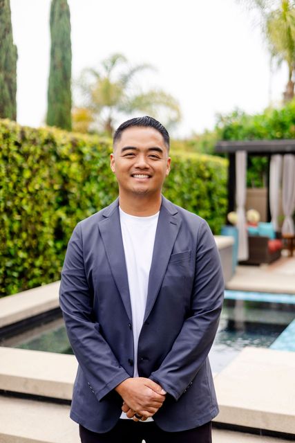 Man standing outdoors by a pool, wearing a dark blazer and white shirt, smiling at the camera.