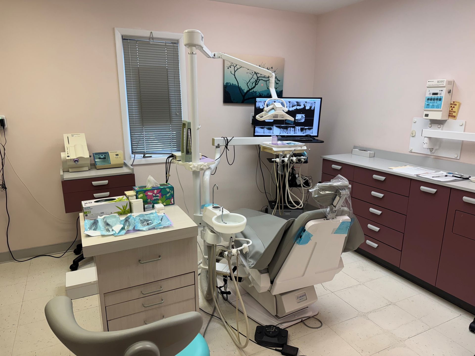 Dental treatment room with chair, monitor, and maroon cabinets, lit by bright overhead lights.