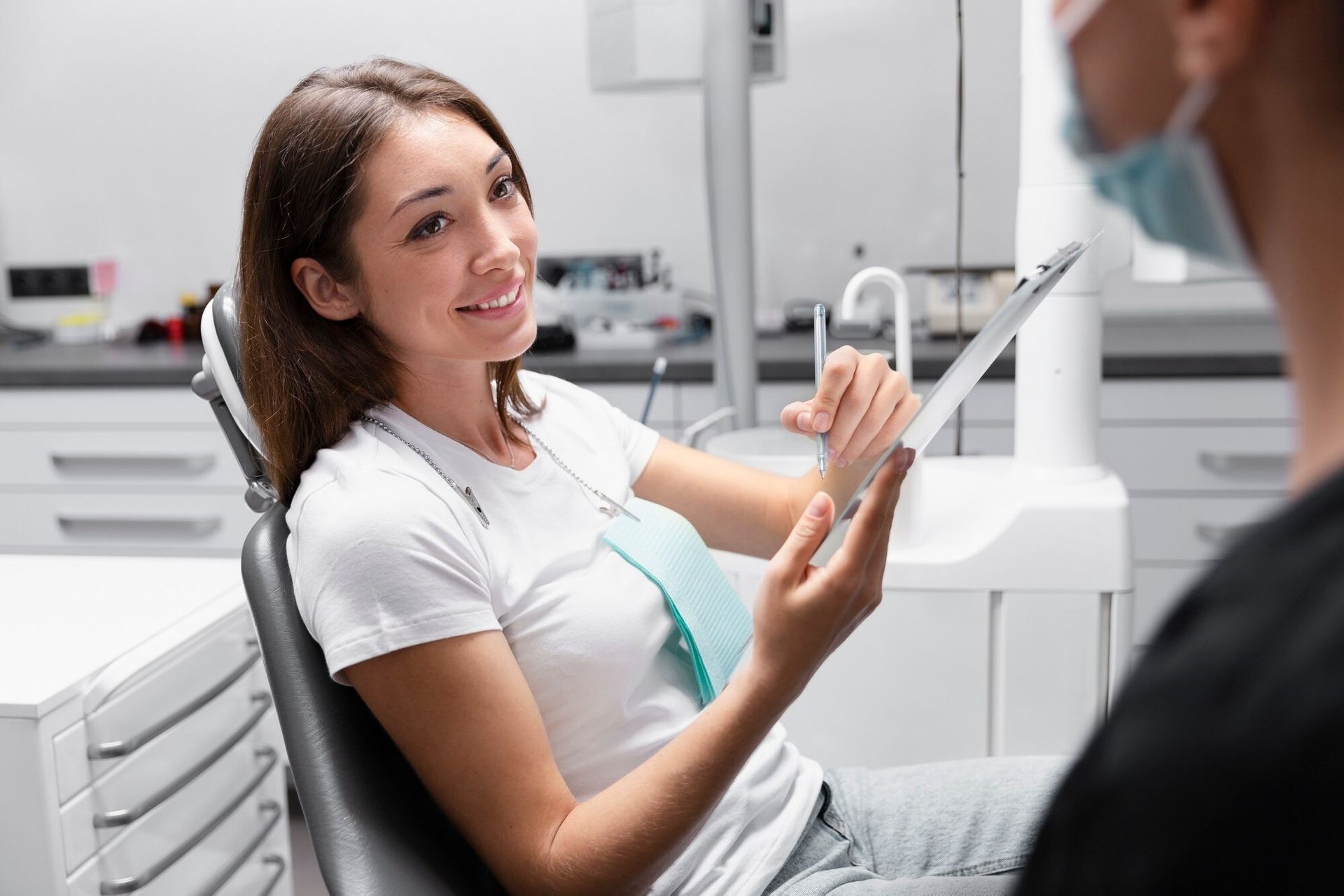 Dental patient smiling while holding a tablet, speaking with a masked dentist in a bright clinic