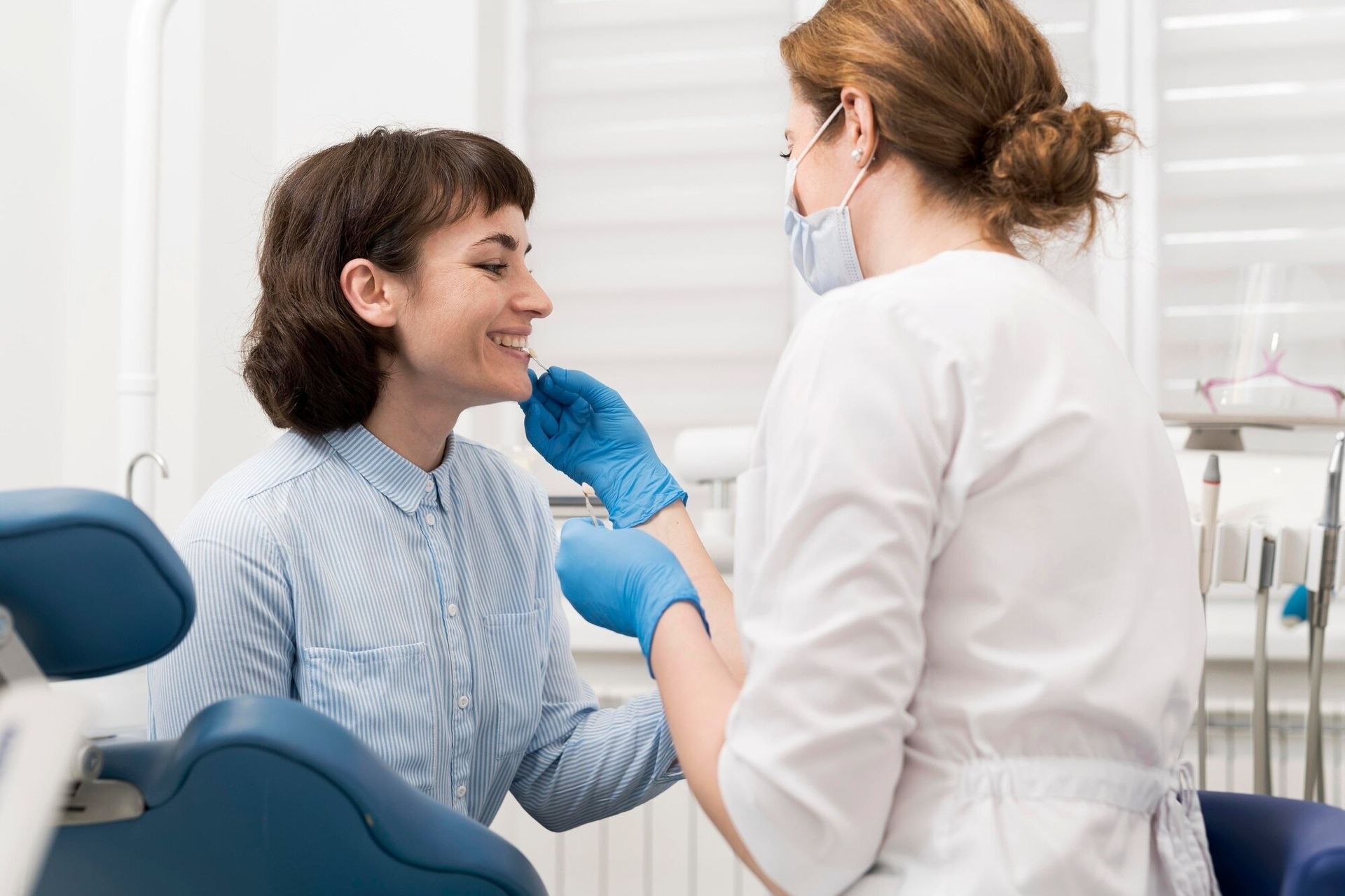 Dentist examining a smiling patient in a bright clinic, wearing blue gloves and a face mask