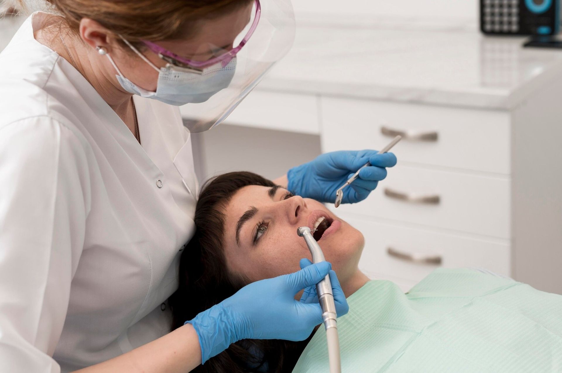Dentist wearing blue gloves examines a patient with dental tools in a bright clinic room