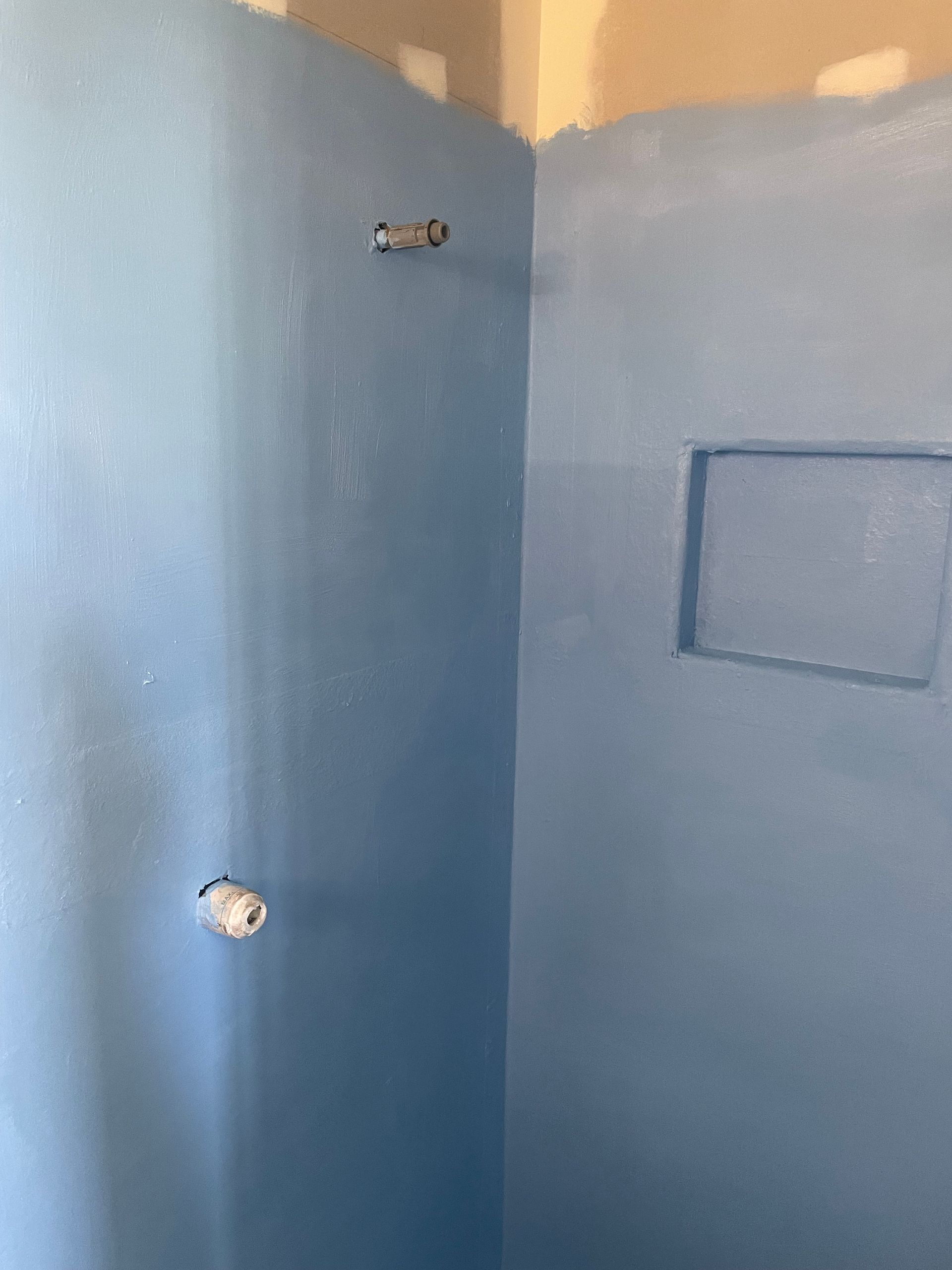 Blue-painted bathroom corner with exposed plumbing and a recessed wall panel.