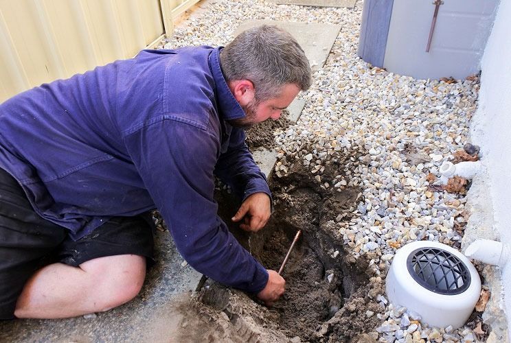 Man kneeling in gravel trench, digging near a white outdoor vent pipe beside a house.