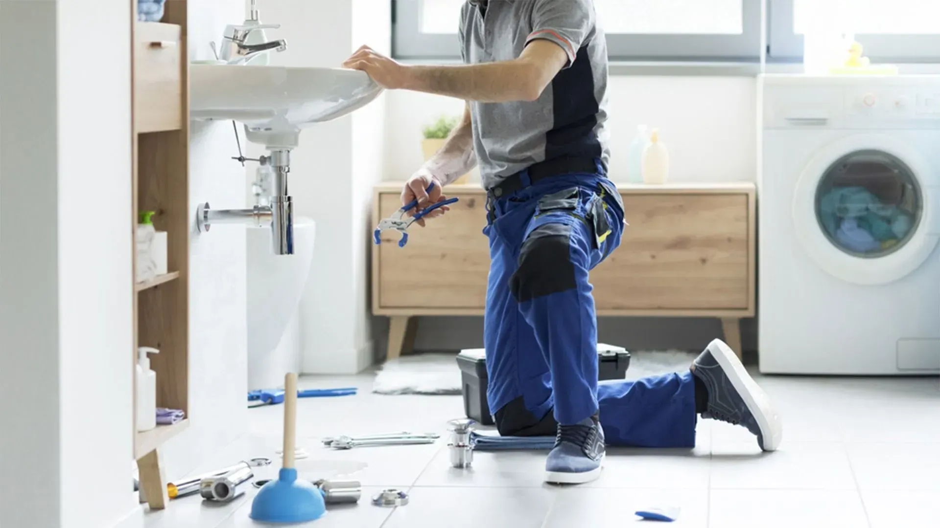 Plumber kneeling under a bathroom sink, fixing pipes in a bright laundry room