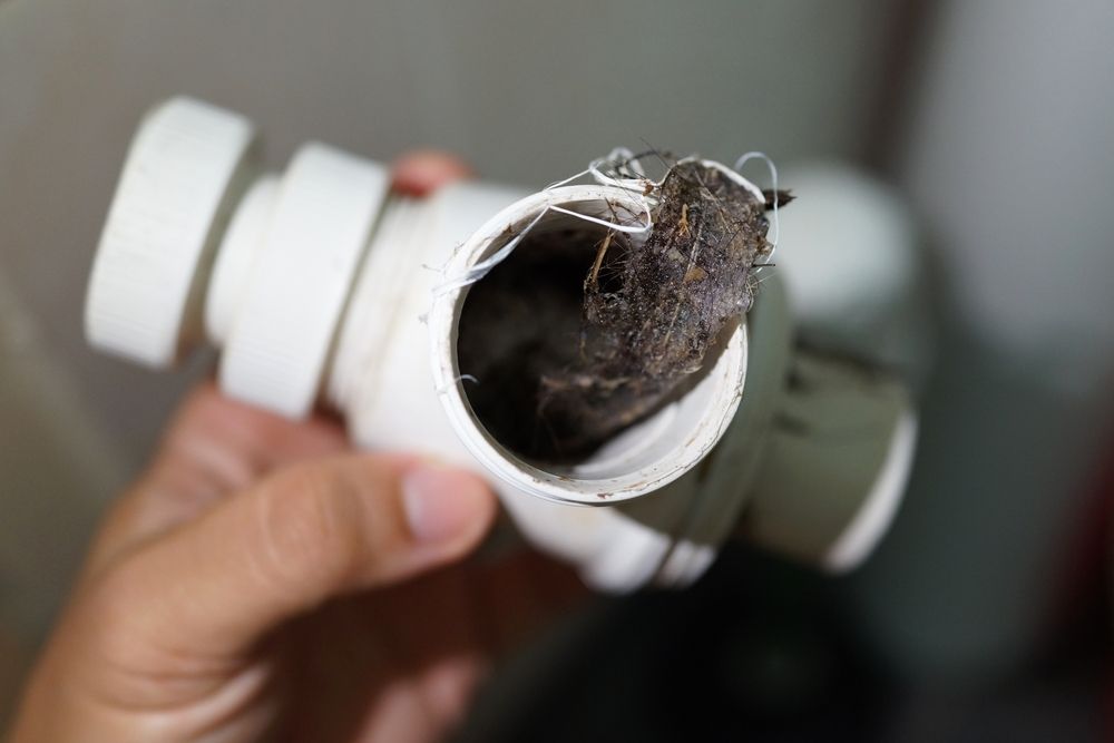 Hand holding a white PVC pipe fitting with a clogged, dirty interior and webbing inside
