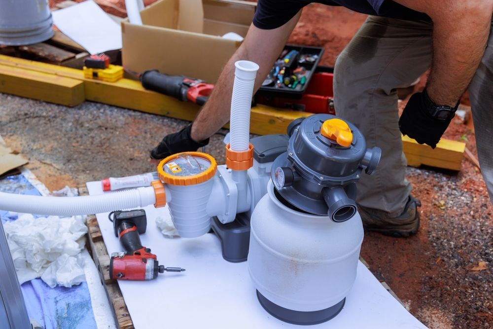 Worker installing white PVC pool plumbing beside a gray filter tank and tools outdoors