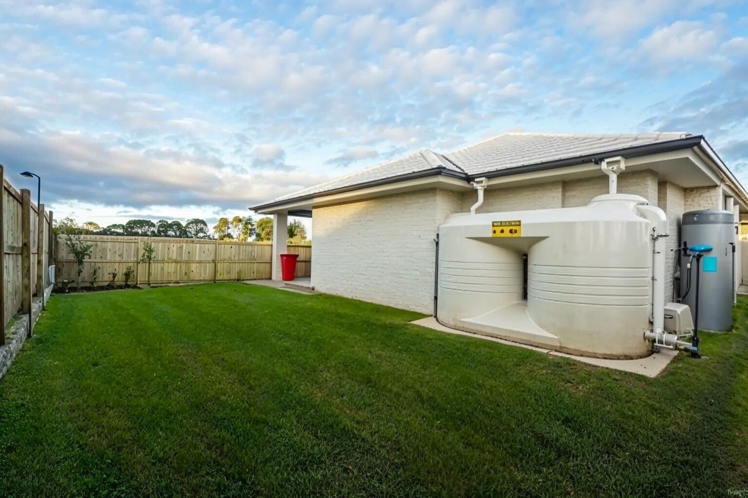 Backyard with green lawn, fenced perimeter, house wall, and large water tanks beside the building