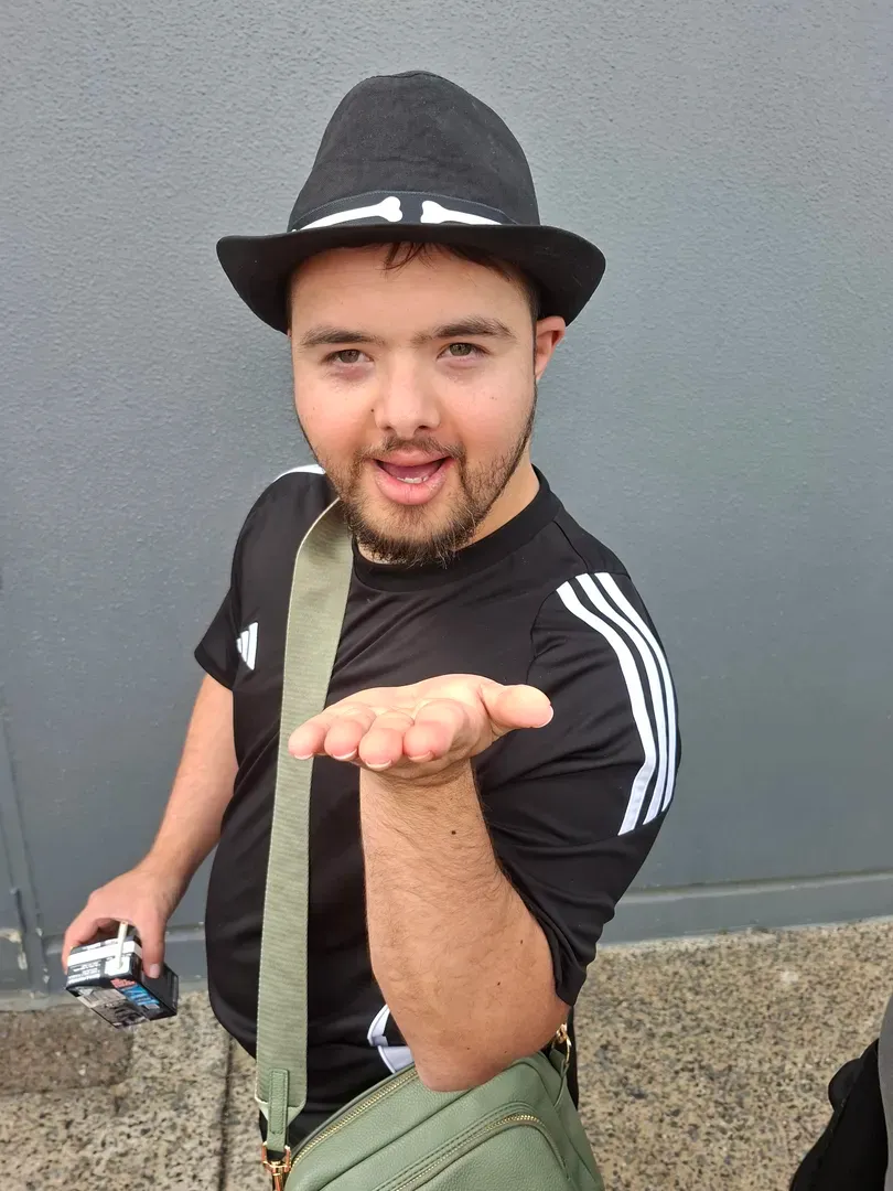 Person in a black hat and shirt blowing a kiss toward the camera, holding a camera against a gray wall.