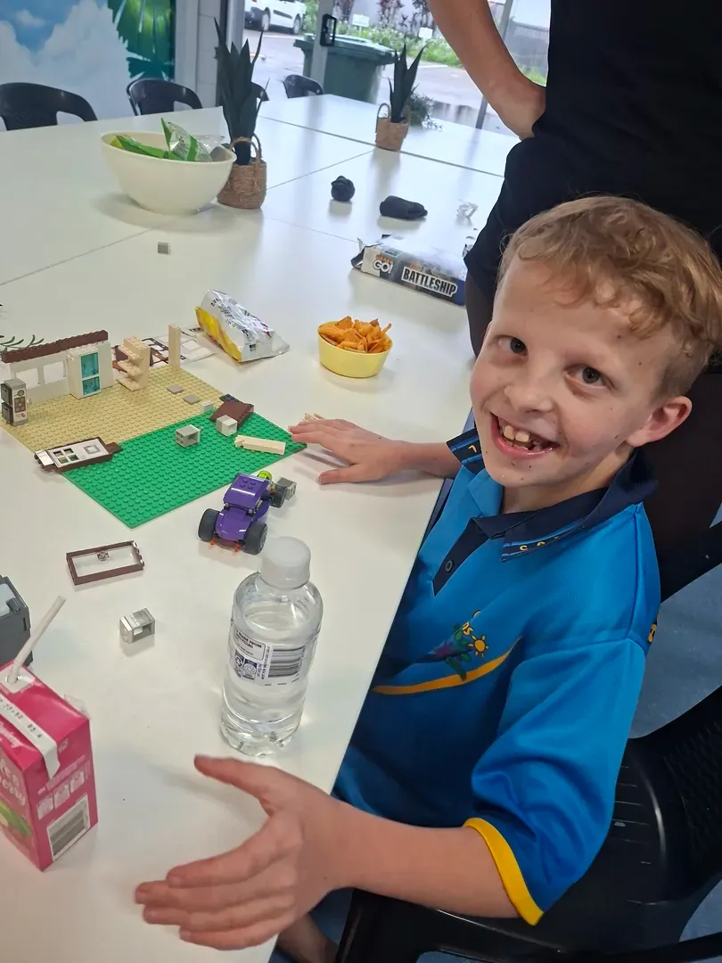 Child smiling at a table with a LEGO setup, water bottle, and toys in a bright indoor space.