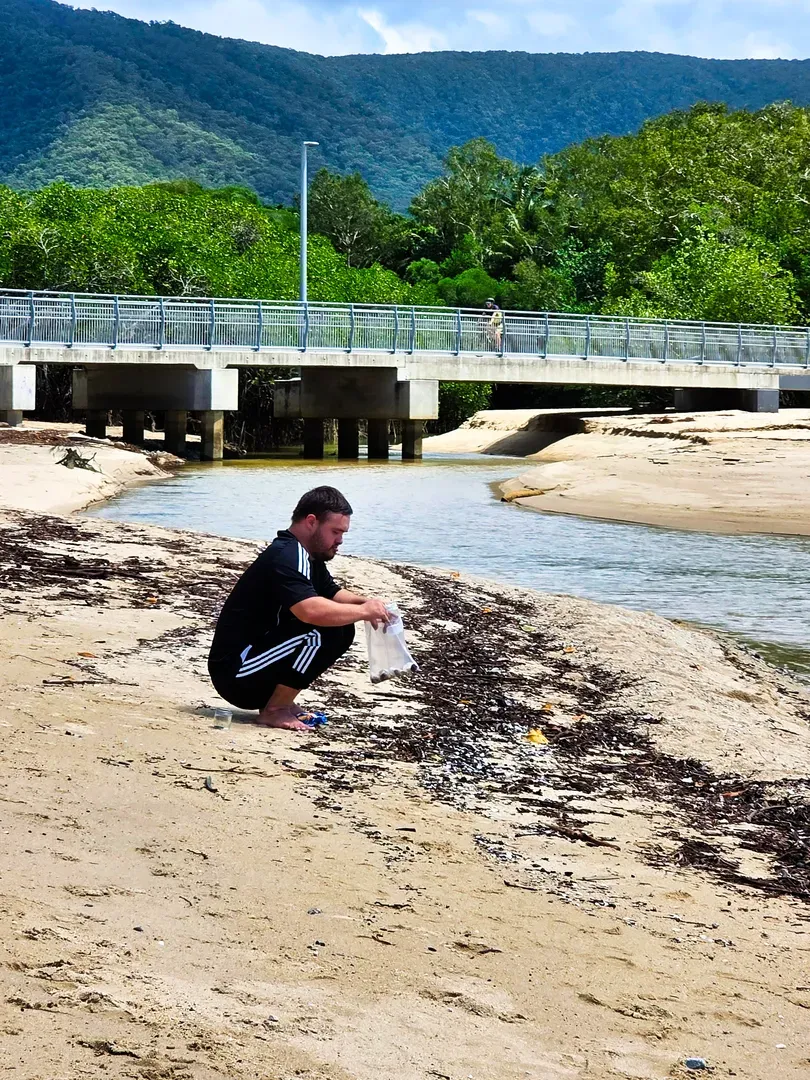 Person squatting on a sandy riverbank beside a shallow stream under a bridge, with green mountains in the background