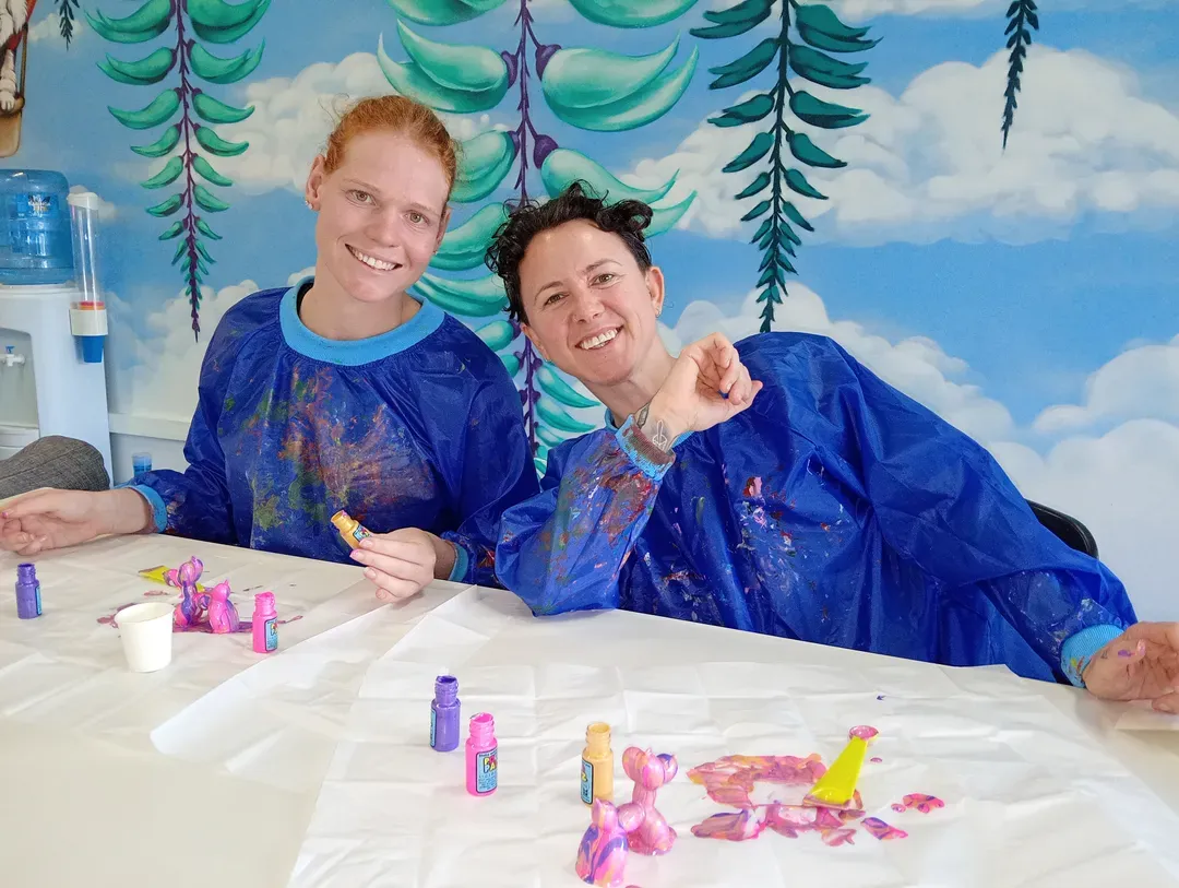 Two people in blue smocks smiling at a paint-covered table in front of a colourful mural