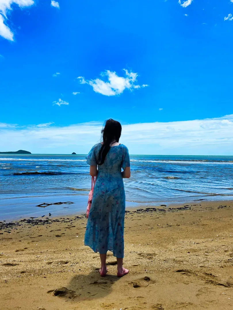 Person standing on a sandy beach facing the ocean under a bright blue sky.