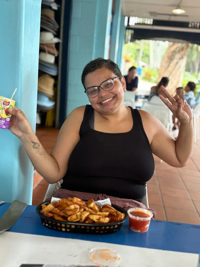 Smiling woman in a black top at a blue table, holding food and a drink in a casual outdoor setting