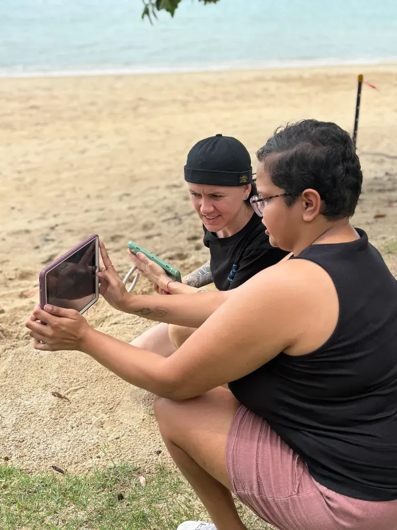 Two people sit on a beach, smiling while looking at a tablet in a phone holder — Sparks Social Abilities in Bungalow, QLD