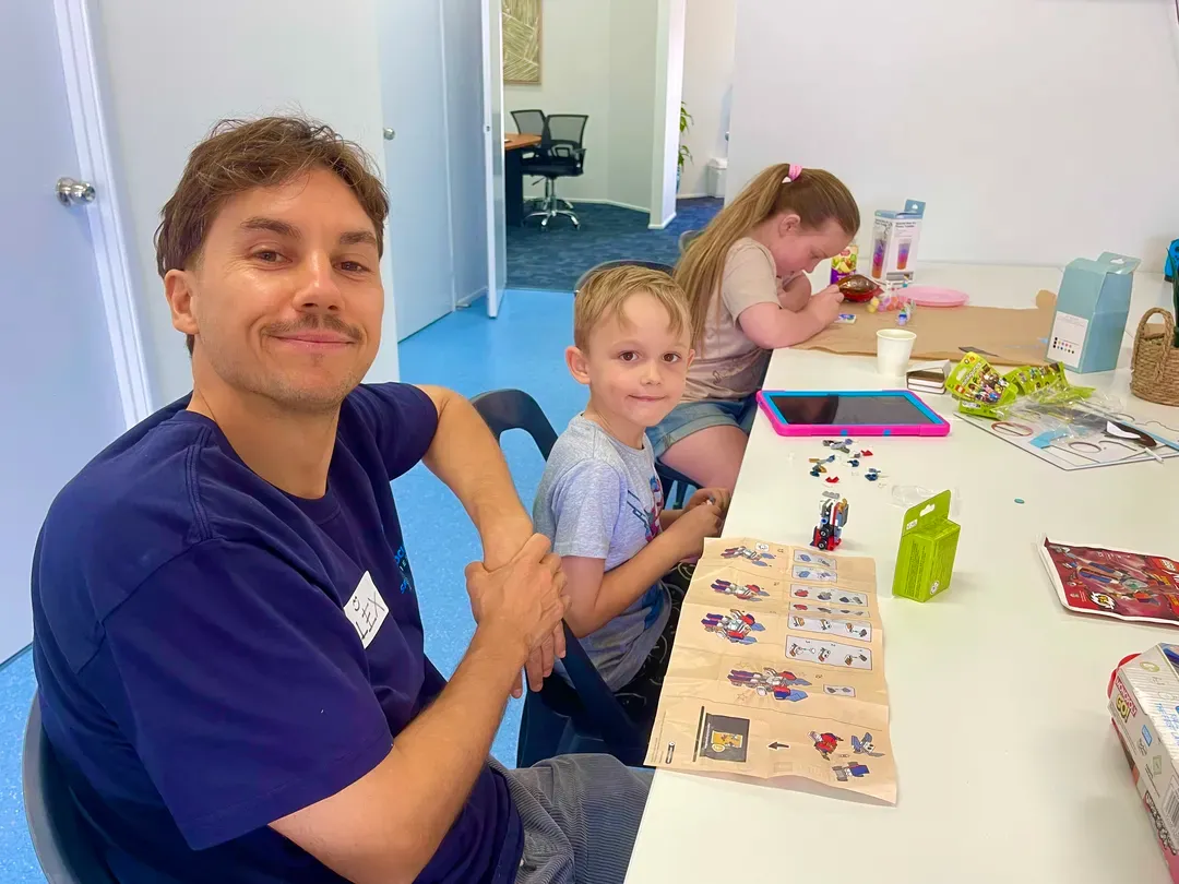 Three people seated at a table, smiling and working on a craft project in a bright room.