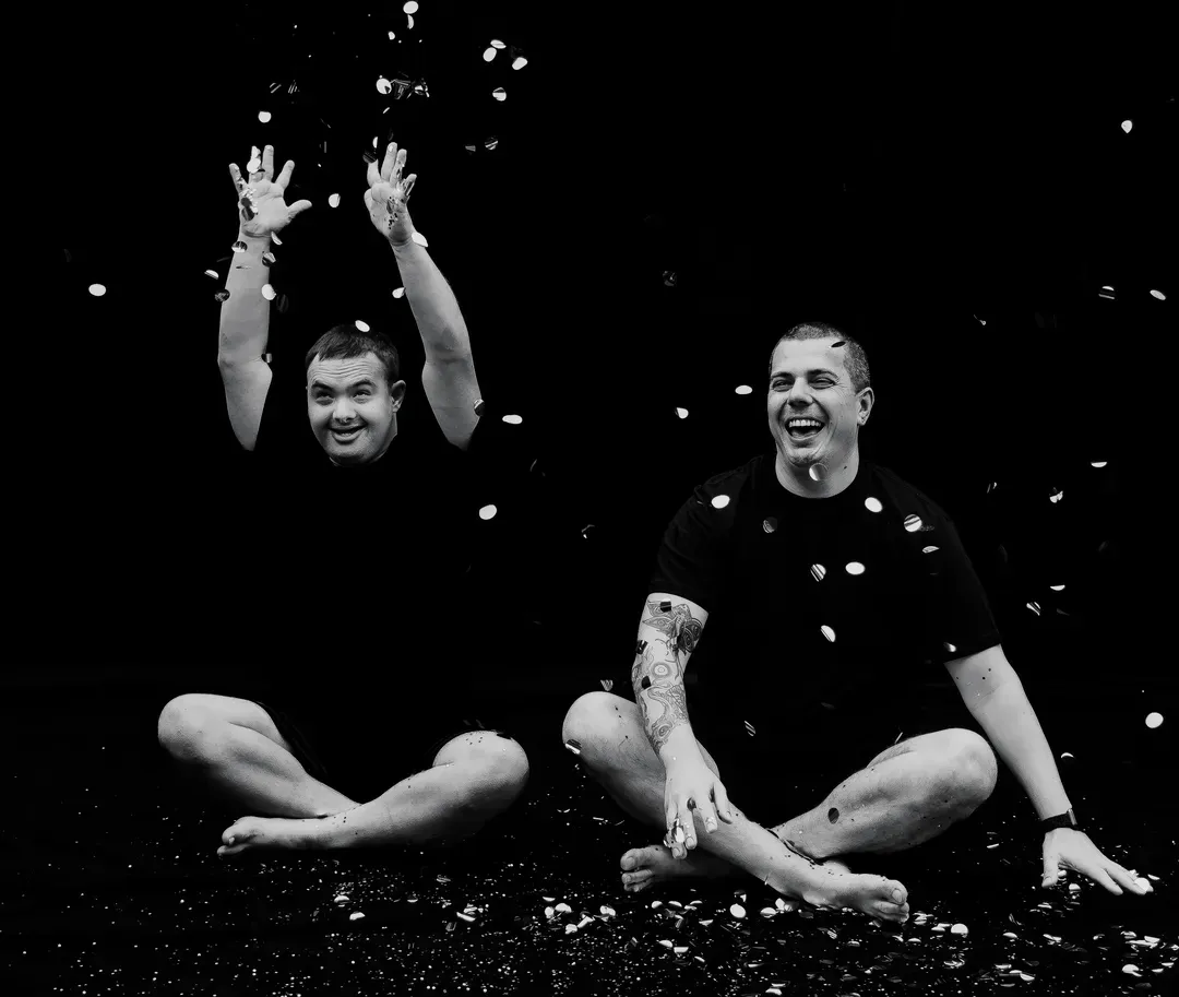 Two people sitting cross-legged and smiling as confetti falls around them on a dark stage.