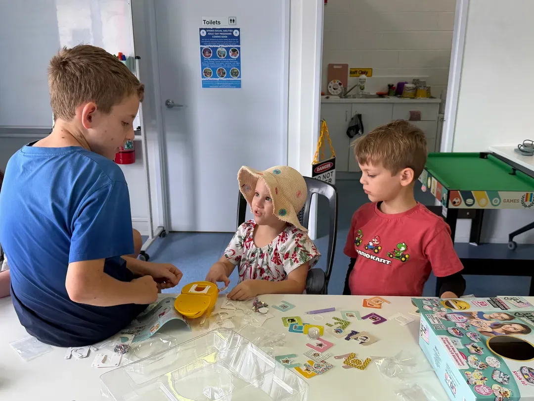 Children sit at a table playing a card game in a bright classroom or playroom.