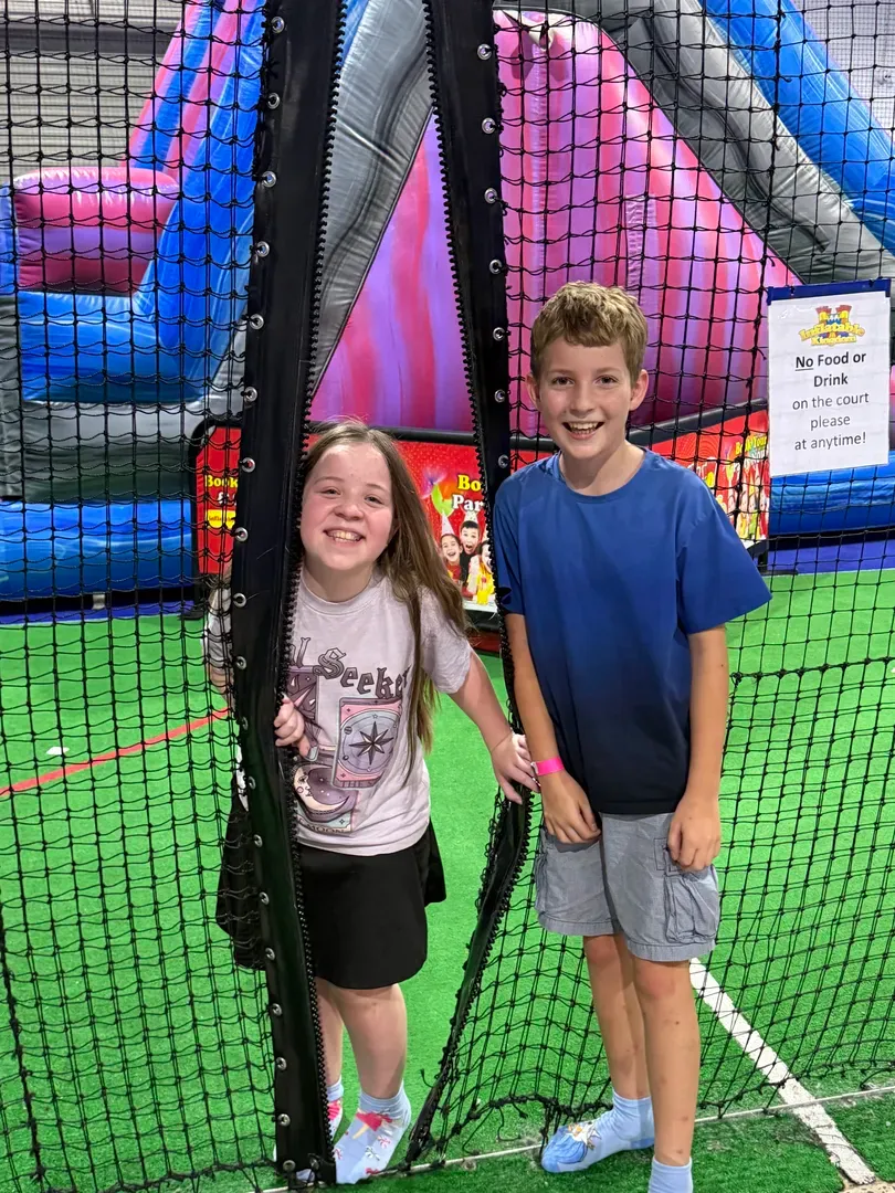 Two children smiling inside an indoor play area with a colourful inflatable slide and netting behind them