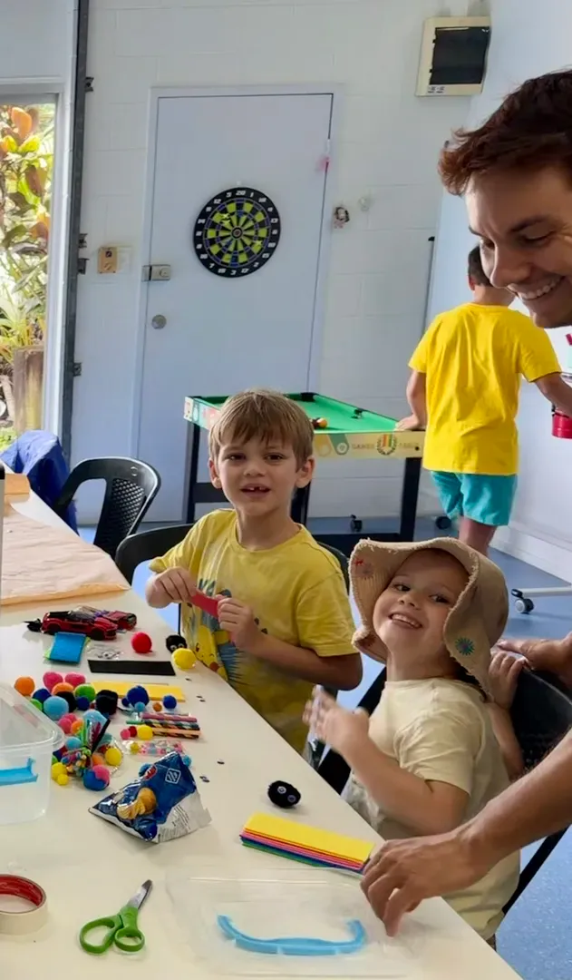 Children making crafts at a table while an adult smiles nearby in a bright classroom.