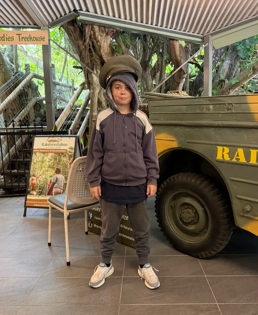 Child standing indoors by a military vehicle, wearing a helmet and gray jacket.