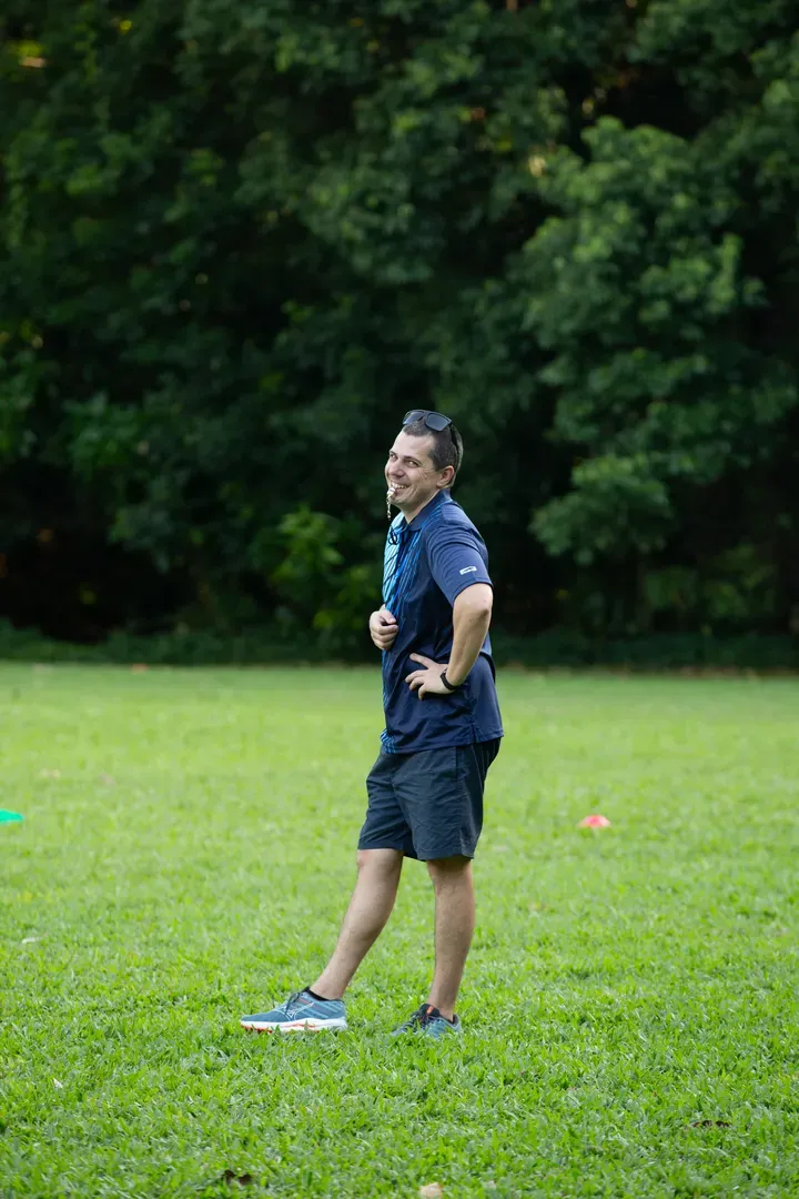 Man standing on a grassy field with hands on hips, trees in the background.