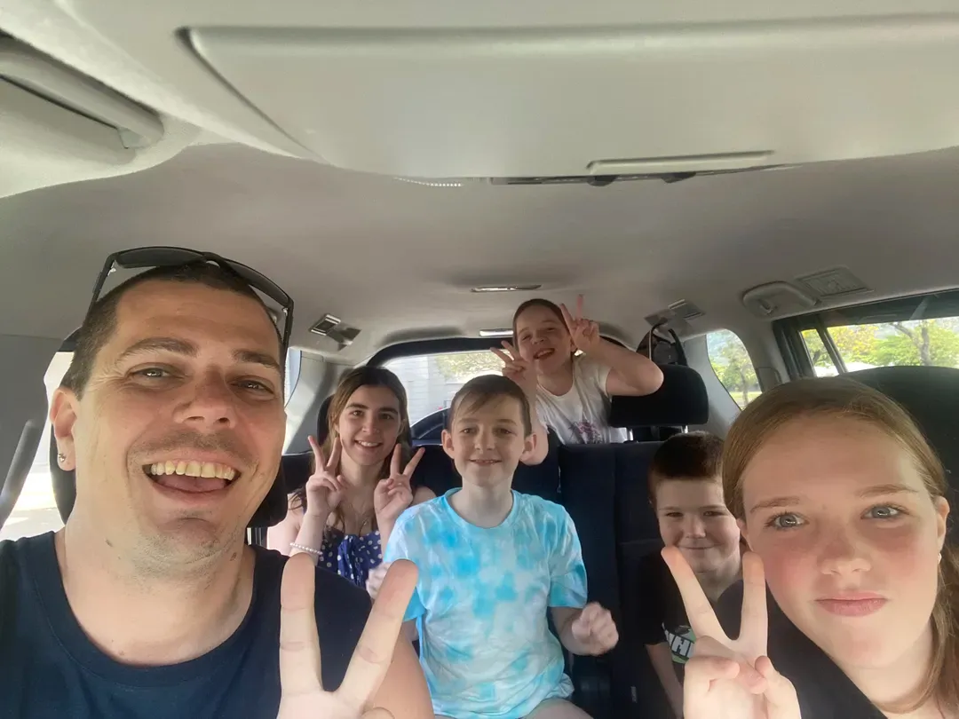 Group of people smiling and making peace signs inside a car, with a child in a blue tie-dye shirt in front.