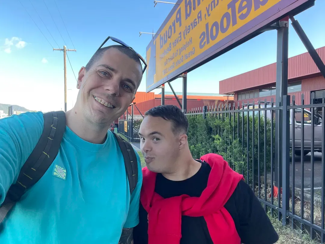 Two smiling people posing outdoors by a metal fence and a yellow sign under a clear blue sky