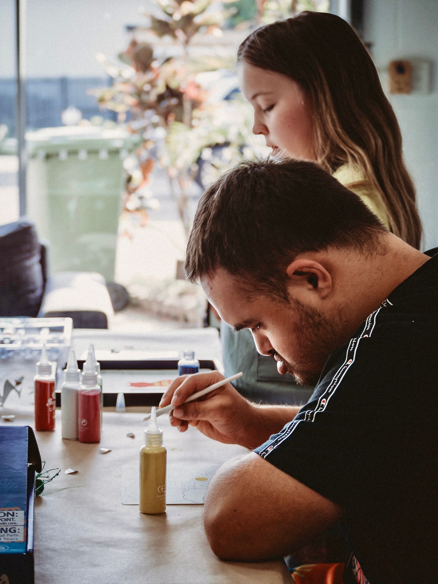 Two people paint small bottles at a table in a bright studio, focused on their work — Sparks Social Abilities in Bungalow, QLD