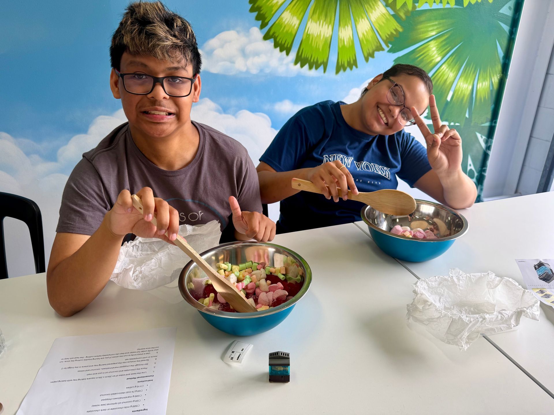 Two people eating from bowls at a table with papers and drinks, under a bright tropical mural.