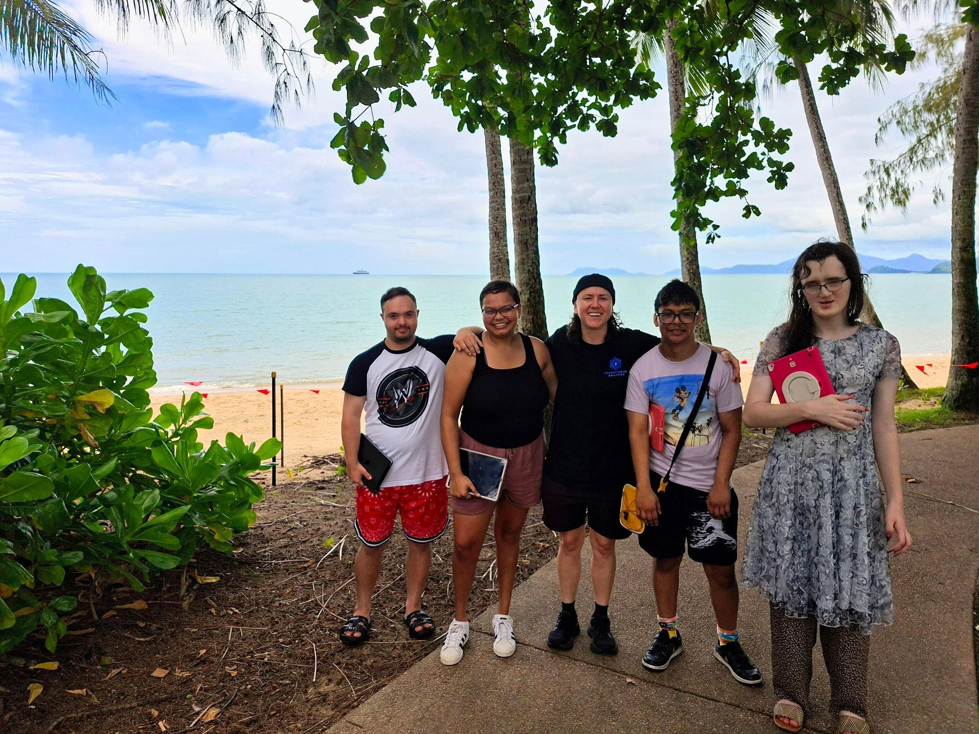 Five people standing on a sandy beach under trees, with ocean and cloudy sky in the background.