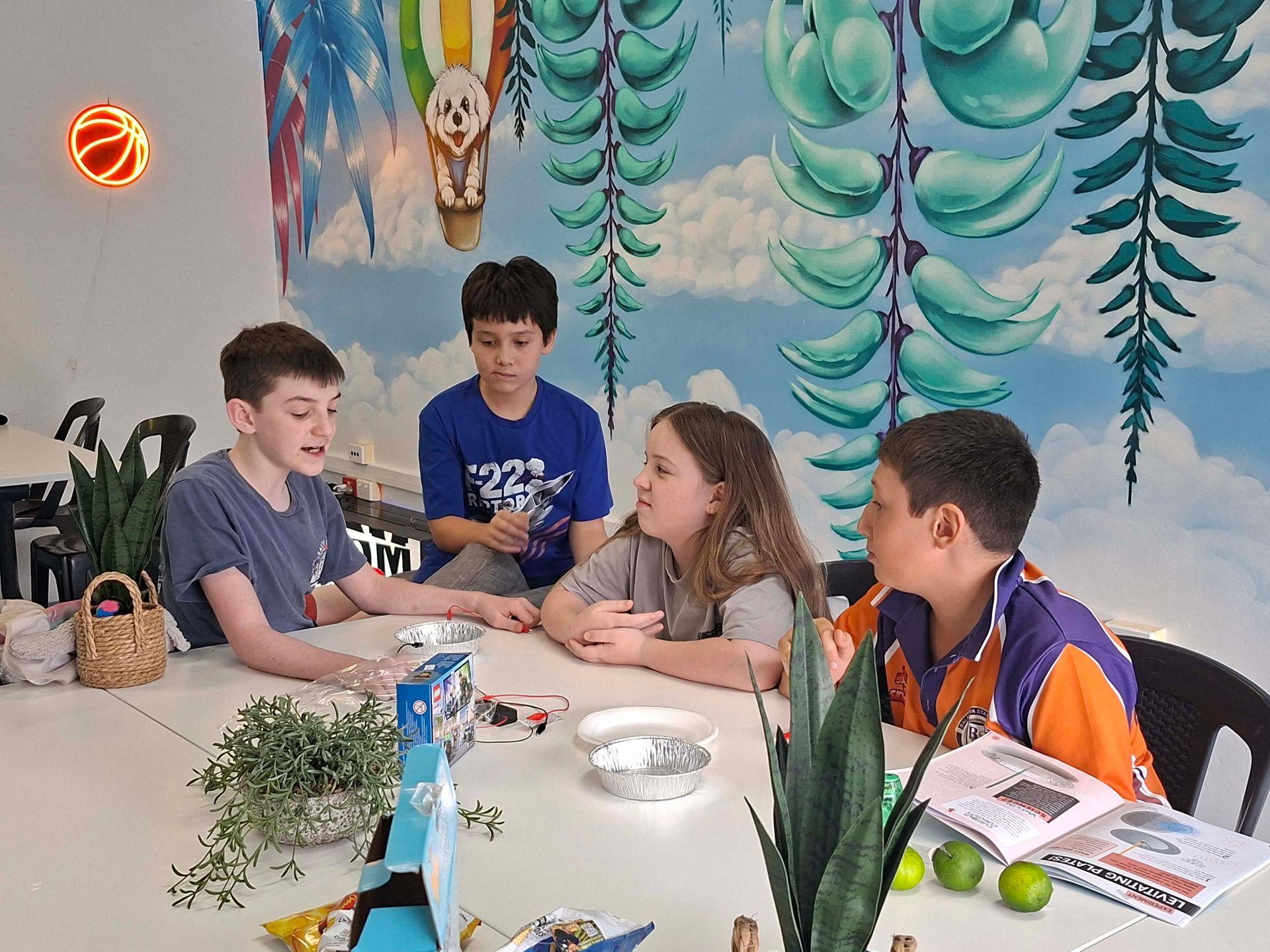 Children sit around a table crafting in a colourful classroom with mural-painted walls and art supplies.