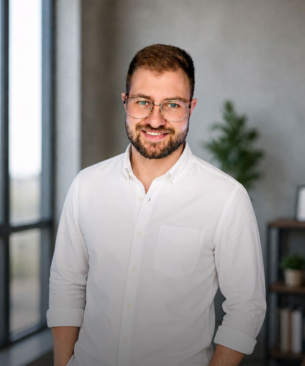 Smiling man in a white shirt and glasses standing indoors, with a blurred plant and gray wall behind him