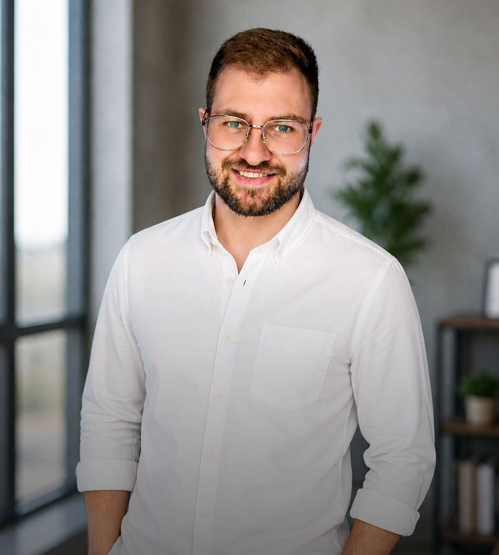 Smiling man in a white shirt and glasses standing indoors with a blurred plant background