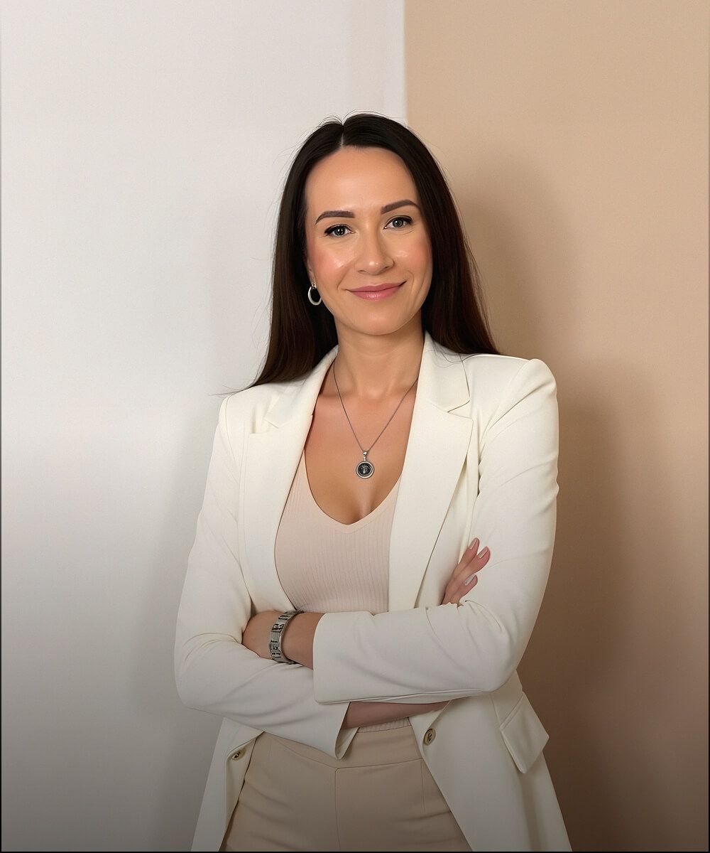 Woman standing with arms crossed, wearing a white blazer and beige outfit against a neutral wall.