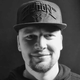 Black-and-white close-up portrait of a man wearing a cap, looking at the camera with a slight smile