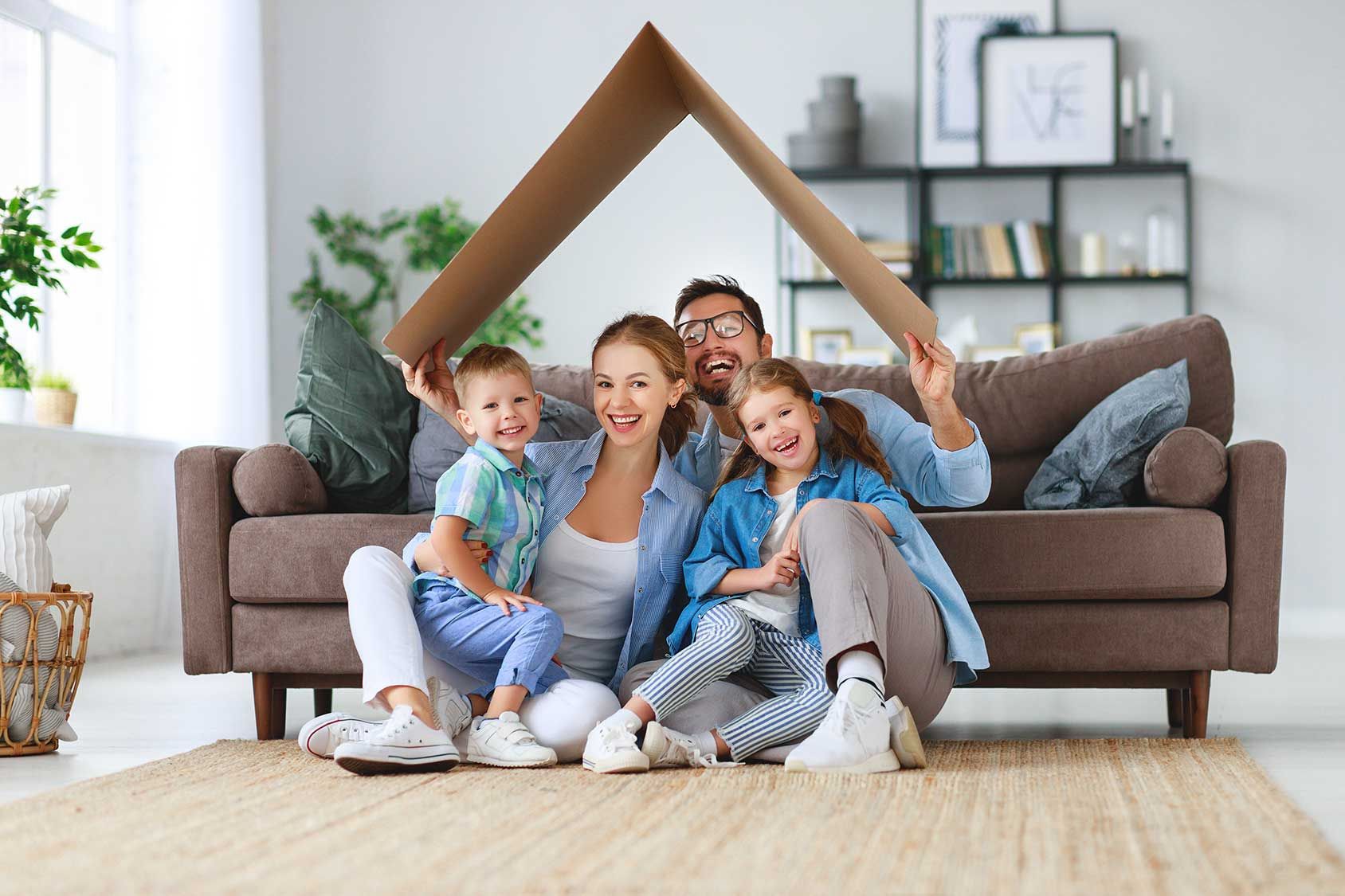Family smiling on a couch in a bright living room, with children holding a roof-shaped cardboard frame