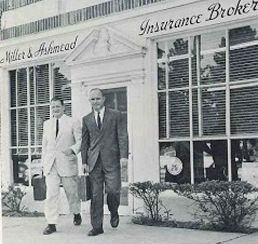 Two men in suits walk outside an insurance broker office with large windows and a sign above the entrance.