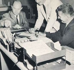 Three men in suits review papers at a cluttered office desk with a typewriter and folders.
