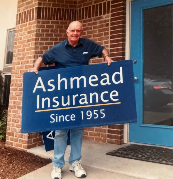 Man standing outside Ashmead Insurance holding a blue sign that reads “Ashmead Insurance Since 1955.”