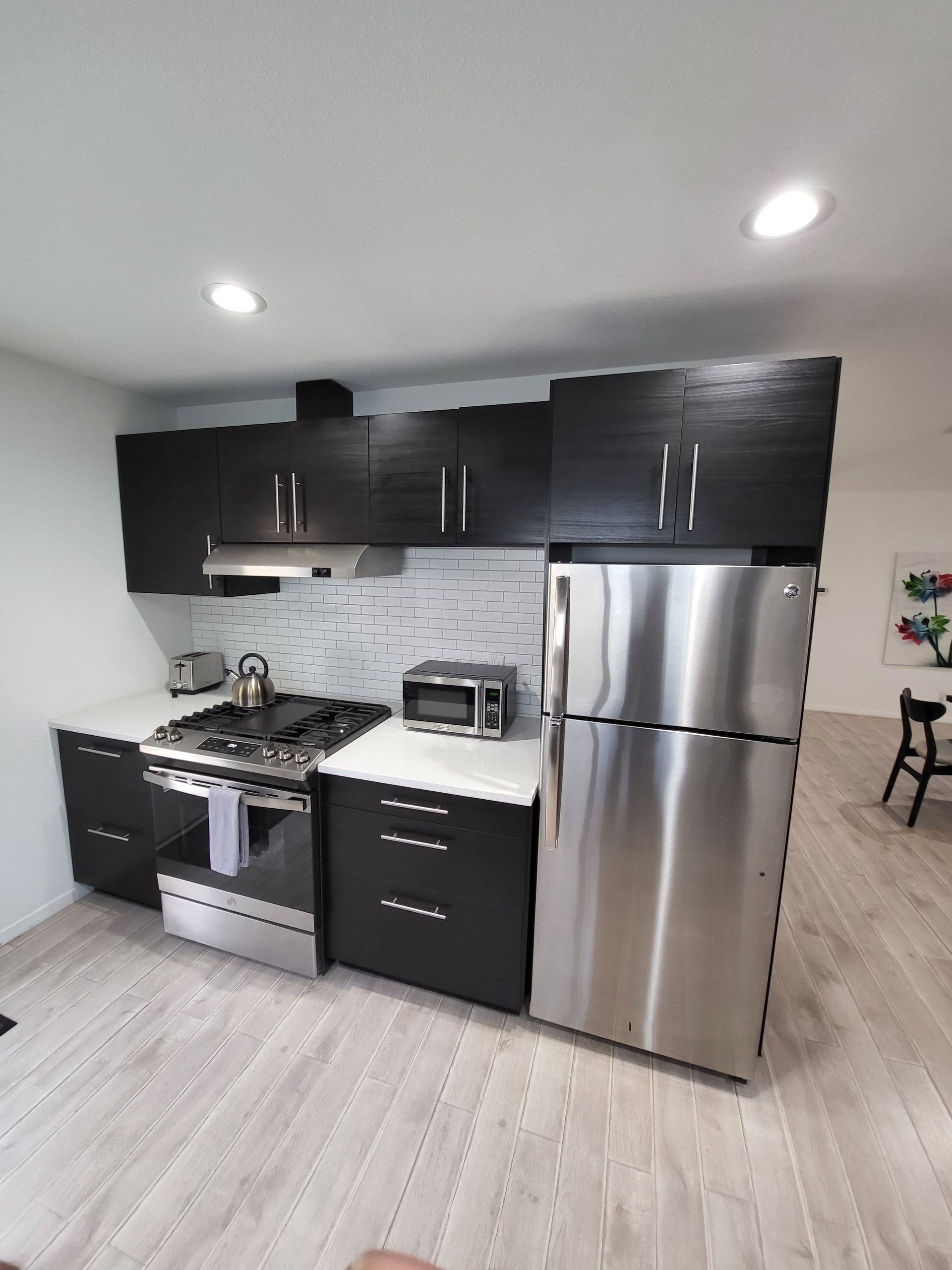 Modern kitchen with dark cabinets, stainless steel appliances, white countertops, and light wood flooring