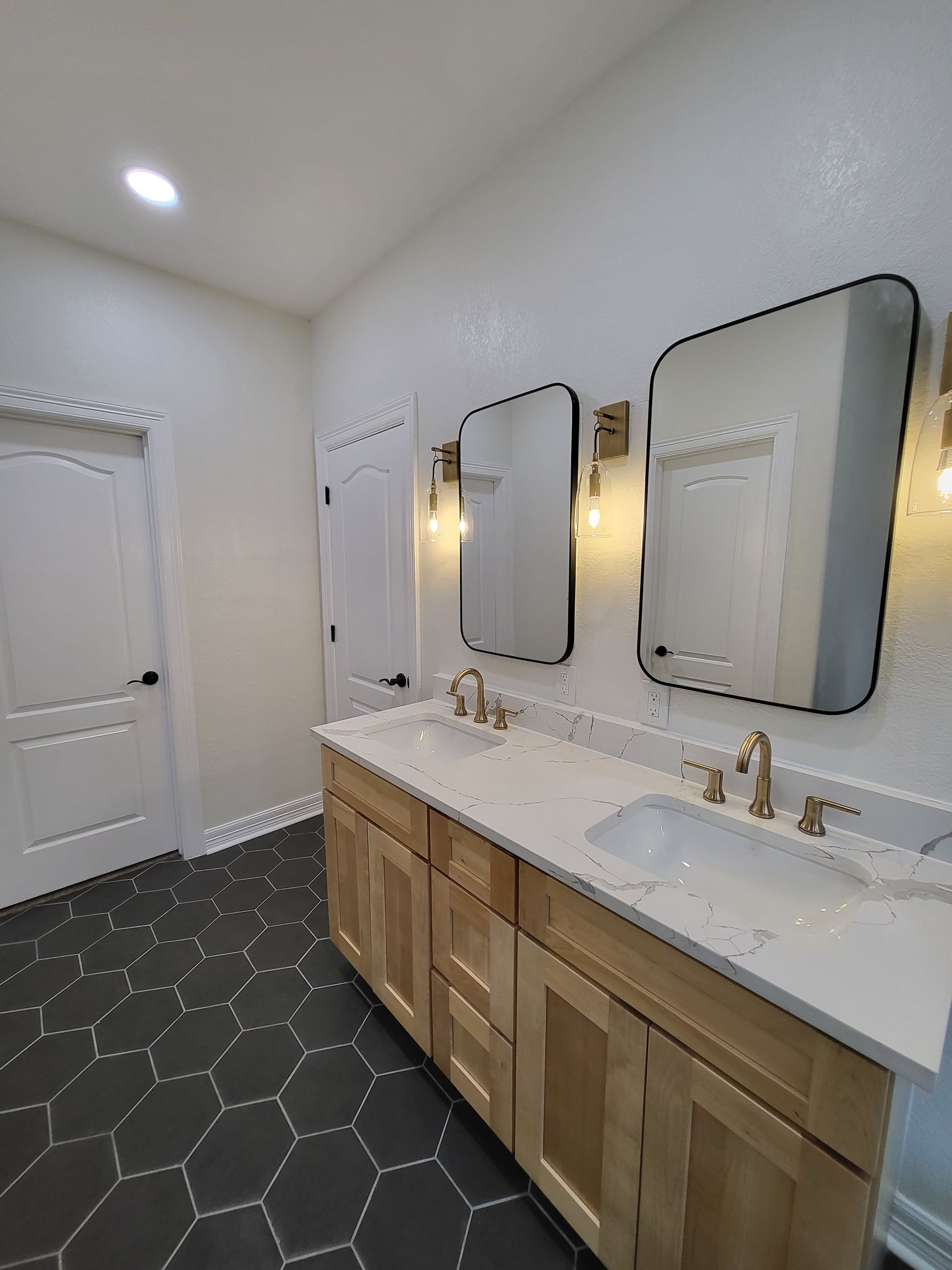 Bathroom with two sinks, two mirrors, gold fixtures, light wood vanity, and dark hexagonal floor tiles.