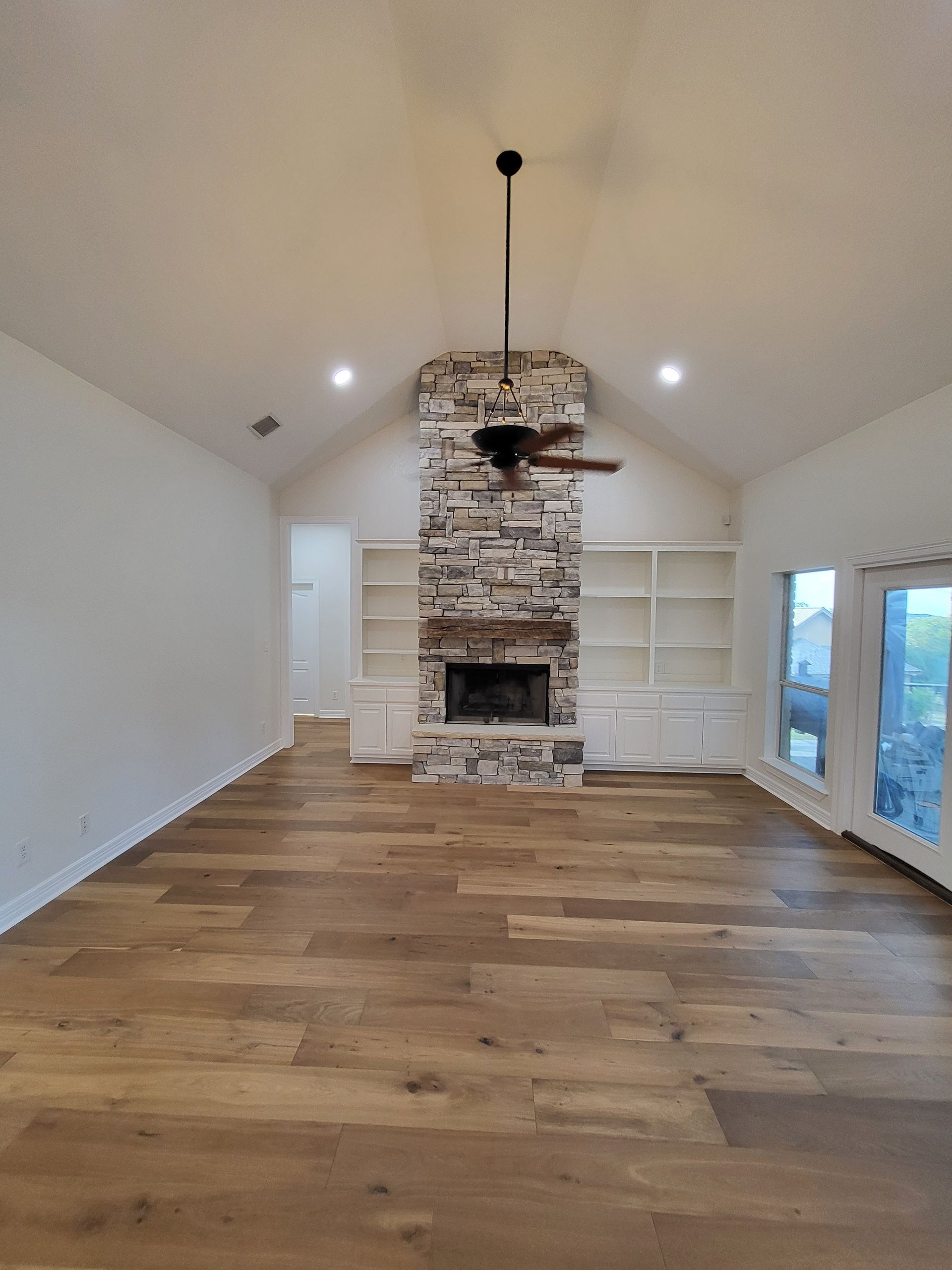 Empty living room with hardwood floors, stone fireplace, built-in shelves, and glass doors to the right.