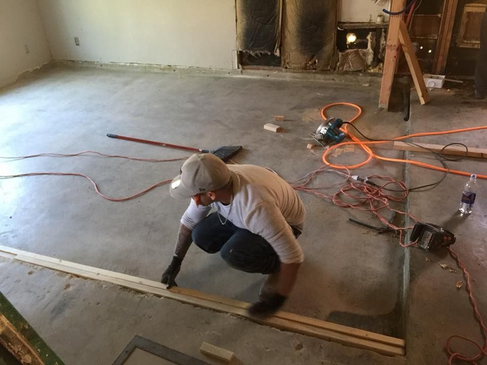 Worker measuring and cutting wood on a concrete floor in an unfinished room with tools and cords nearby