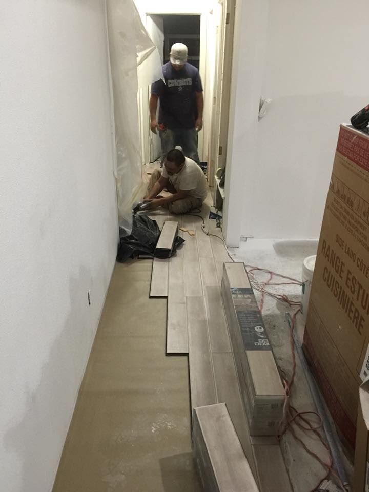 Two workers installing floor tiles in a narrow hallway with tools and materials scattered around.