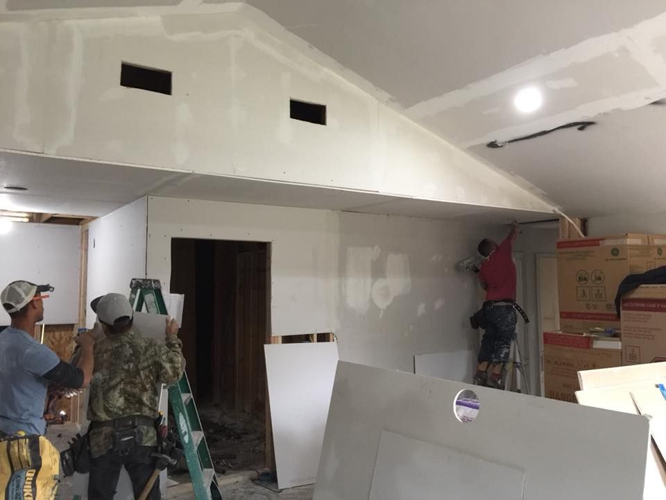 Workers painting drywall inside a room under construction, with ladders and materials stacked nearby.